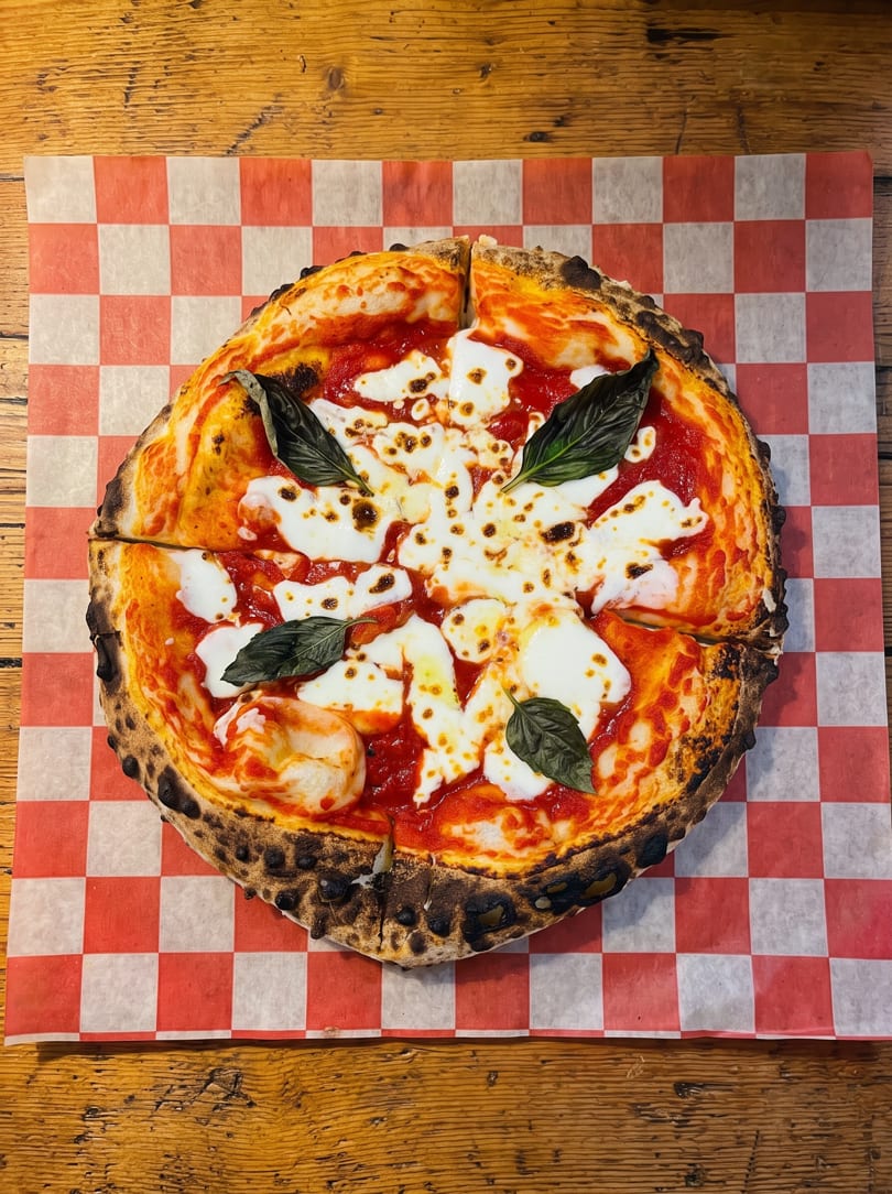 Neapolitan-style Margherita pizza with charred crust, fresh mozzarella, and basil on checkered paper.