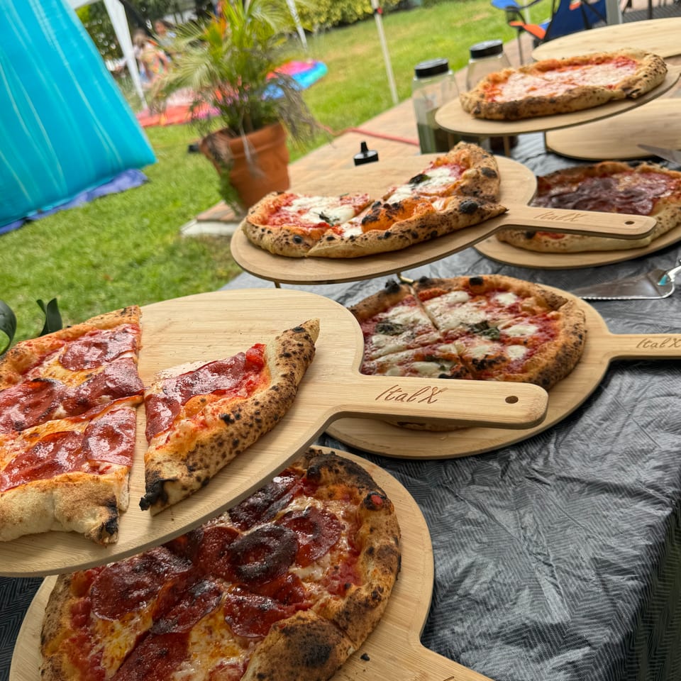 Freshly baked pepperoni and Margherita pizzas on wooden ItalX peels at an outdoor party.