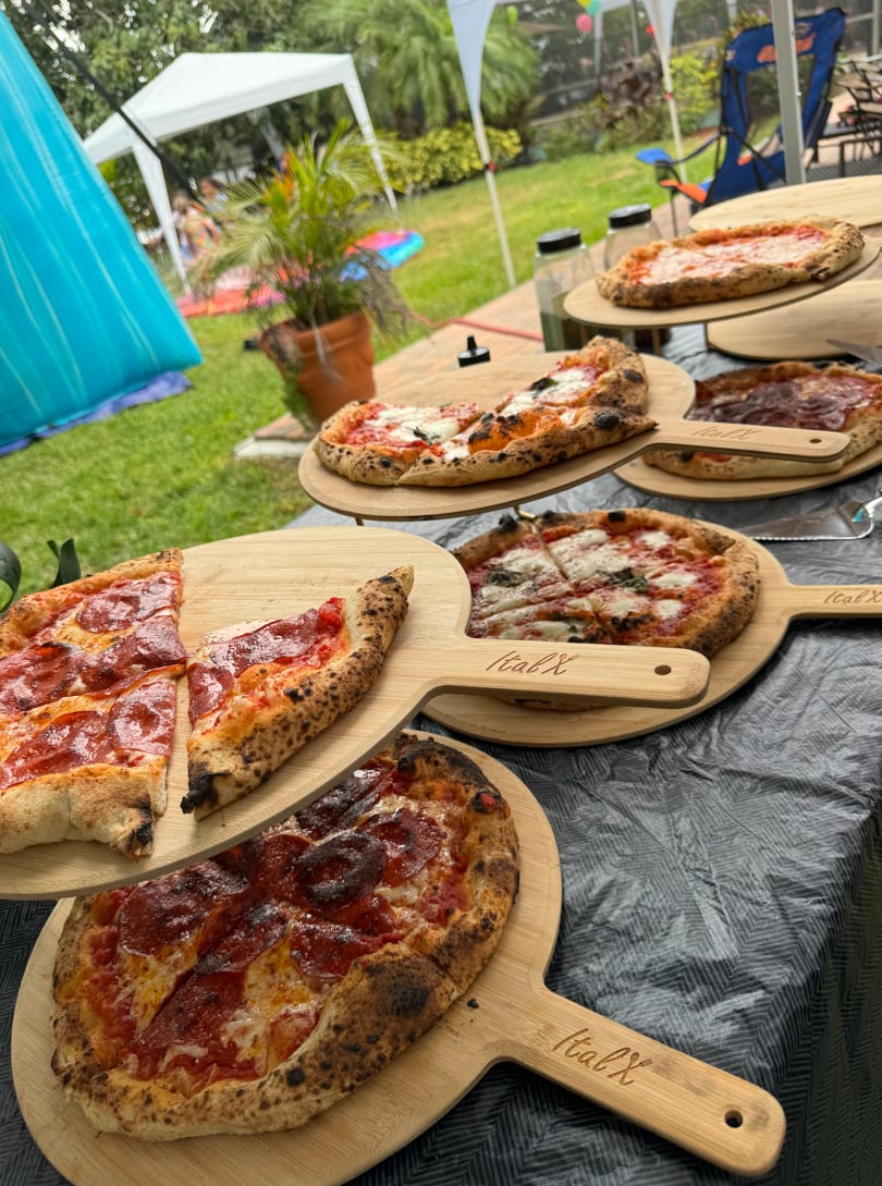 Freshly baked pepperoni and Margherita pizzas on wooden ItalX peels at an outdoor party.