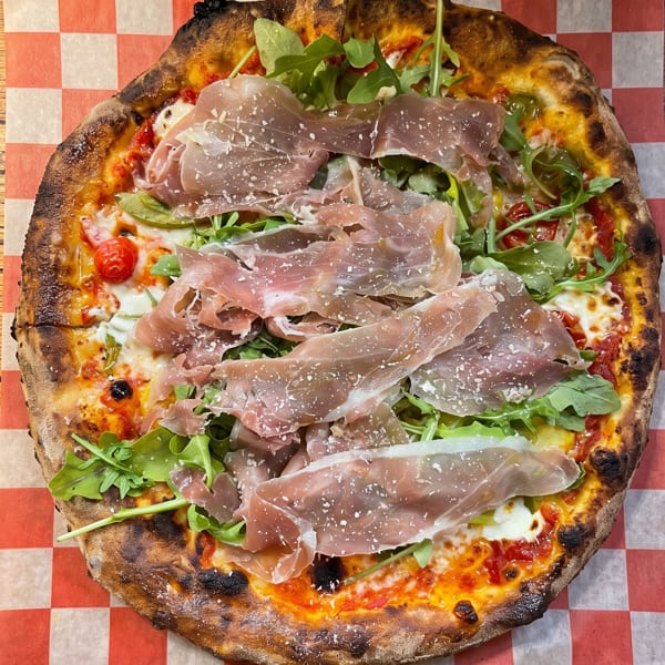 Wood-fired pizza with prosciutto, arugula, cherry tomatoes, and parmesan on checkered paper.