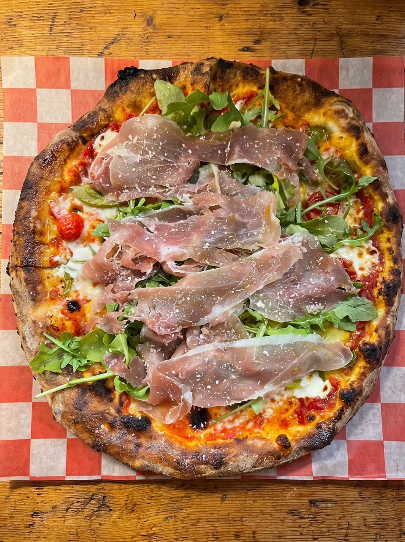 Neapolitan pizza with prosciutto, fresh arugula, cherry tomatoes, and cheese on checkered paper.