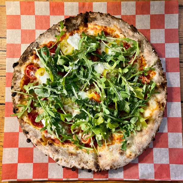 A whole pizza with arugula, burrata, cherry tomatoes, and banana peppers on a checkered paper.