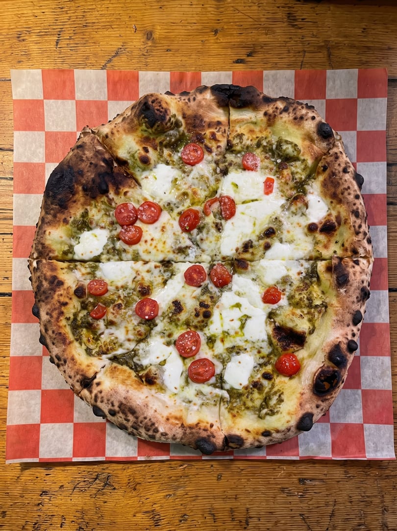 Pesto pizza with mozzarella, sliced cherry tomatoes, and a charred crust on checkered paper.